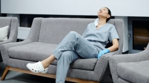 Νοσοκόμα καθισμέη σε καναπέ με ρούχα εργασίας και σαμπό